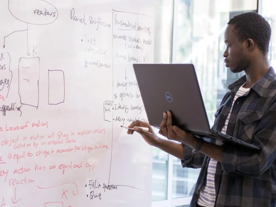 A student holds a laptop and writes on a dry erase board.