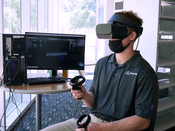 A student sitting next to a computer monitor and wearing a VR headset.