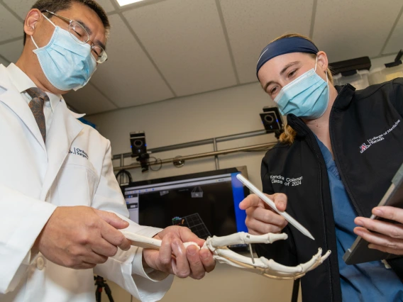 A professor and a student, both wearing surgical masks, seen from below. They are looking down at a model of the hand and wrist bones.