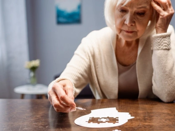 An older woman sitting at a table completing a white puzzle shaped like a human head.