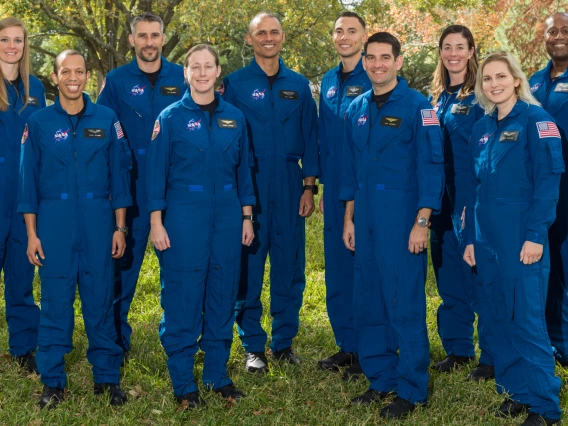 A group of ten astronauts stand in matching blue jumpsuits in a green outdoor area.