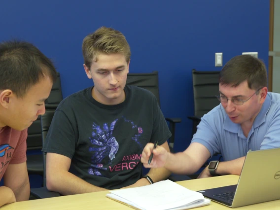 Three men lean over a laptop and set of papers on a table.