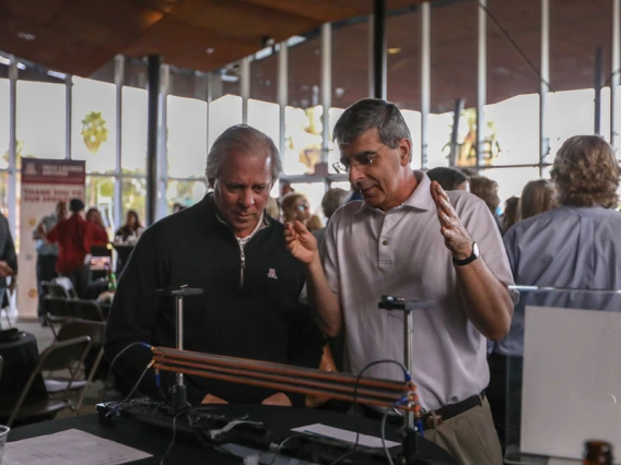 Two men stand in a crowded lobby. One is gesturing at machine on a table in front of him, while the other listens carefully.