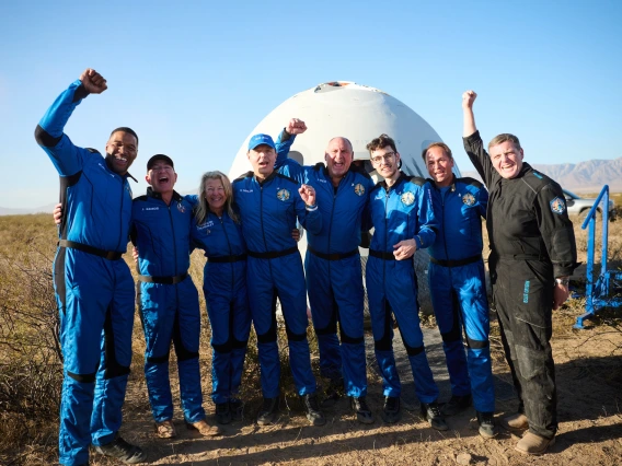 A group of eight people in blue jumpsuits smile in front of a round spacecraft. They look happy, with their arms around each other or raised in the air in celebration.