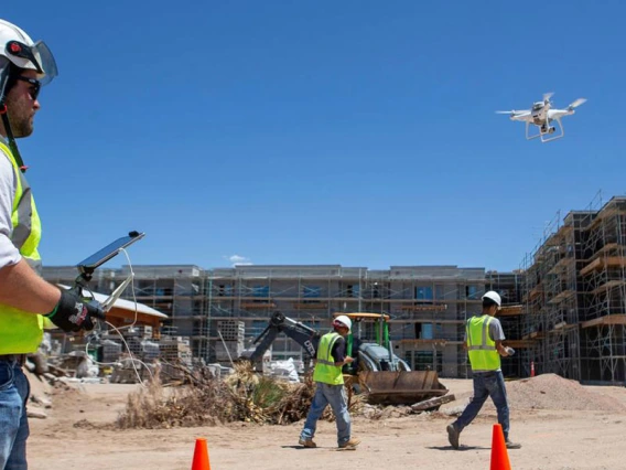 A man in a yellow construction vest and hard hat operates a drone on a construction site.