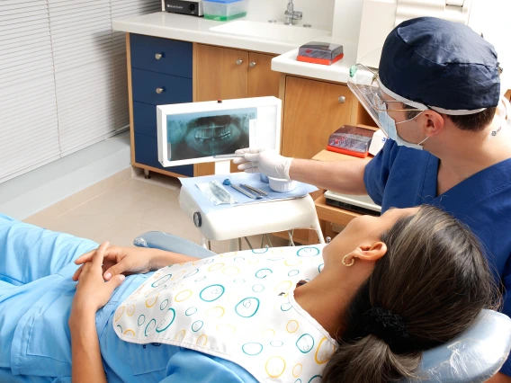 A dental patient lies in a chair while a dentist gestures toward X-rays of the patient's teeth.