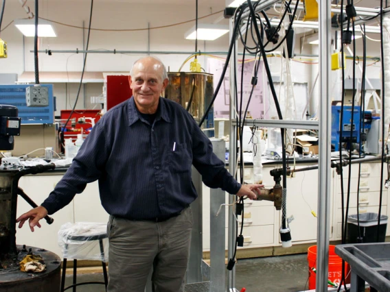 Don Gervasio, wearing a long-sleeved blue shirt, stands between a pump and pipe in a laboratory with tubes hanging in the background