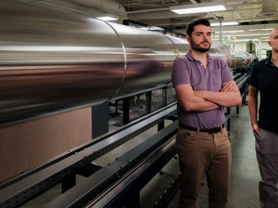 Alex Craig and Jesse Little stand next to LT5, a long wind tunnel which is silver and stretches off frame to the left of them.