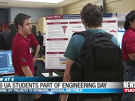 A student in a red polo stands in front of a scientific poster talking to someone in a green shirt reading the poster.