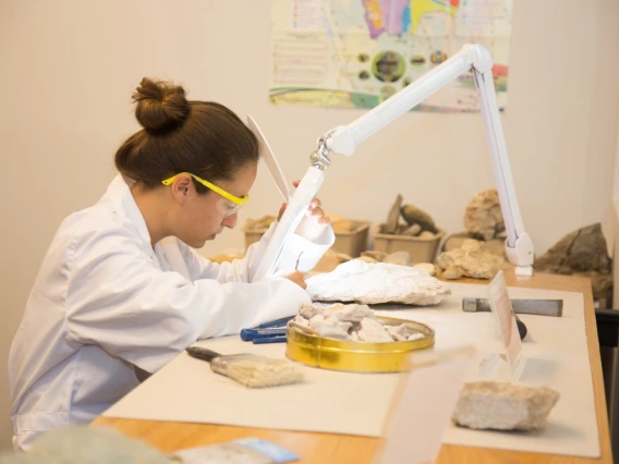 A young woman in a lab coat and goggles leans over a sample of rocks laid out on a table. They are lit up with a bright light shining from a flexible arm.