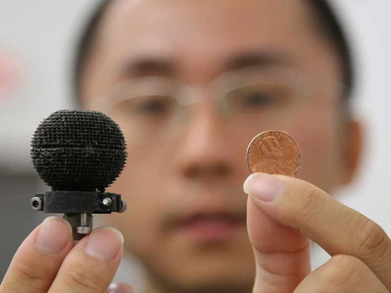 A man holds up a small 3D-printed black sphere in one hand, a penny in the other. The sphere is only about 3 times the size of the penny.