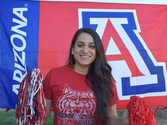 Maira Garcia smiles and holds two pom poms. Behind her is the University of Arizona Block A logo.