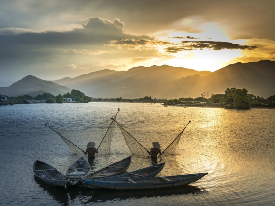 two fishers standing in water and holding up nets. There are four canoes behind them. In the distance is a sunset over mountains.