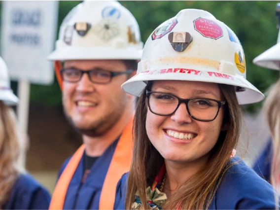 A student smiling and wearing graduation garb and a white hard hat covered in stickers. There are three other students in the frame, wearing the same combination, but out of focus.