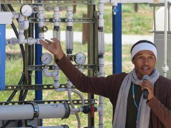 A young man holding a microphone gestures toward a systems of pipes and gauges.