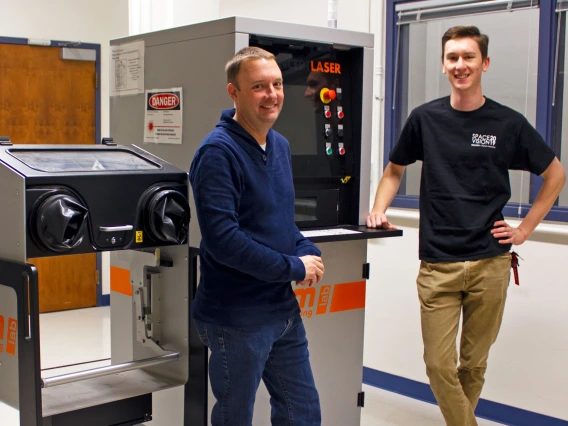 Two young men standing next to a machine about the same height as them, with a screen and a series of buttons. It says "LASER" at the top.