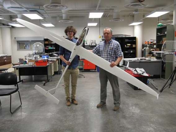 Two men stand in a large indoor space with concrete floors, holding a white sailplane about 7 feet long and with a 12 foot wingspan.