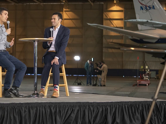 Two men sit on a stage with a NASA plane in the background