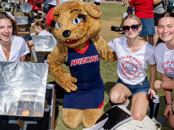 Three students and the Wilma Wildcat mascot pose outdoors with a solar oven