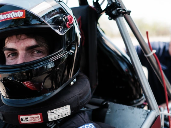 A close up photo of a student wearing a dark motorcycle helmet and sitting in a Formula-style vehicle. A man is behind him working on the vehicle.