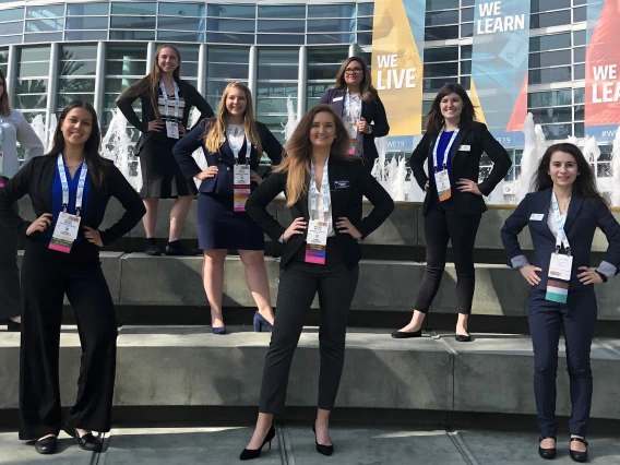 Eight women in professional attire stand on three levels of steps posing for a photo in front of the Anaheim Convention Center.