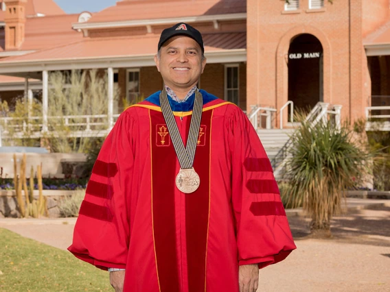 Ricardo Valerdi, wearing academic regalia and a medal, poses in front of Old Main on the University of Arizona campus