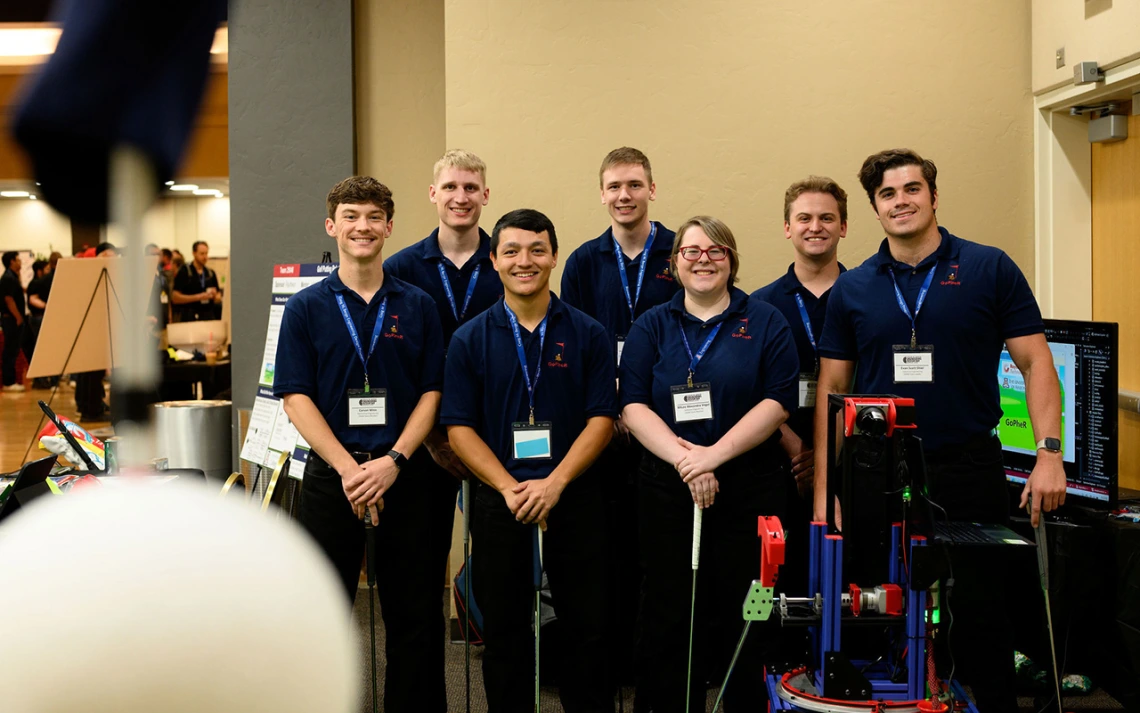 Six men and one woman pose for a photo next to a golfing robot.