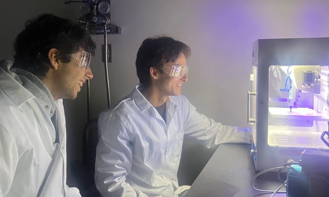 Two students with dark hair look at a 3D printer.