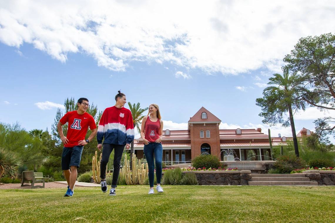 Three students in red shirts walk on a green lawn.