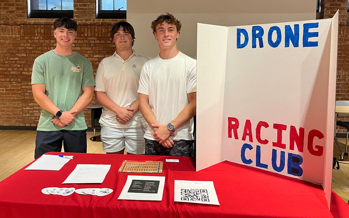 Three young men stand behind a red table with a poster board that reads: Drone Racing Club.