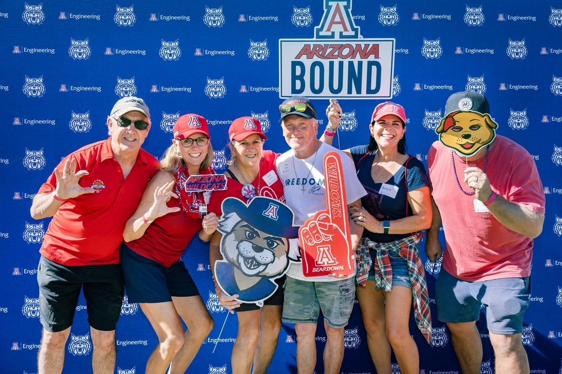 Six adults in multicolored shirts stand in front of a U of A College of Engineering photo wall.