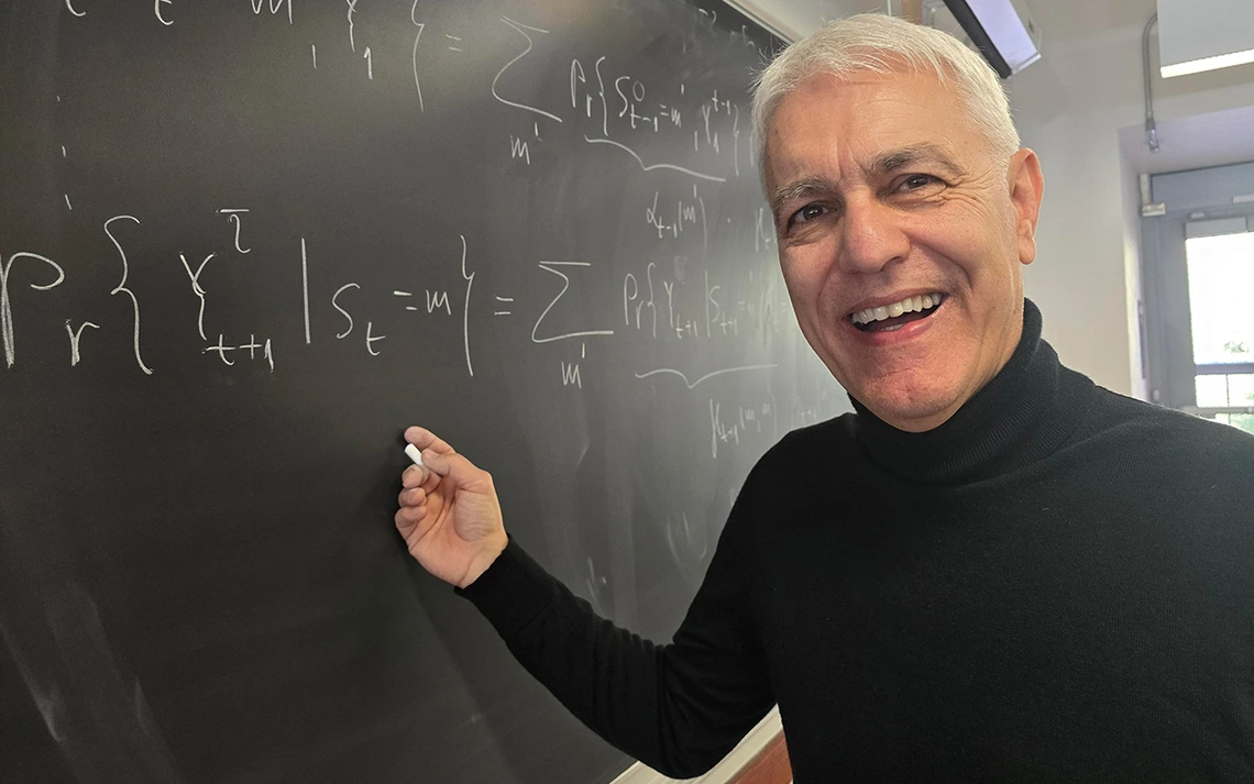 A man in a black turtleneck sweater stands in front of a black chalkboard.