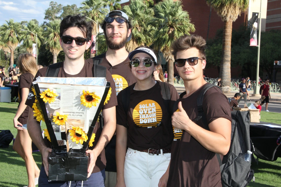 Four students gather around their solar oven, which is made of cardboard and foil and decorated with plastic sunflowers