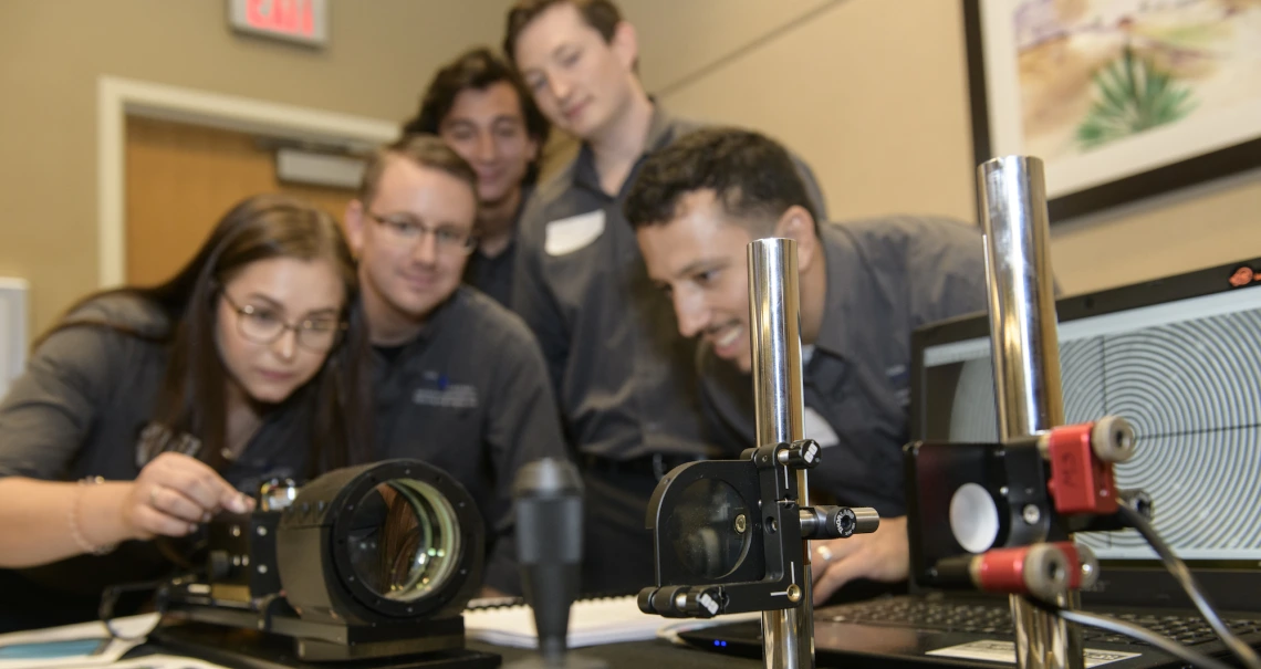 Five students lean over a display of optical equipment