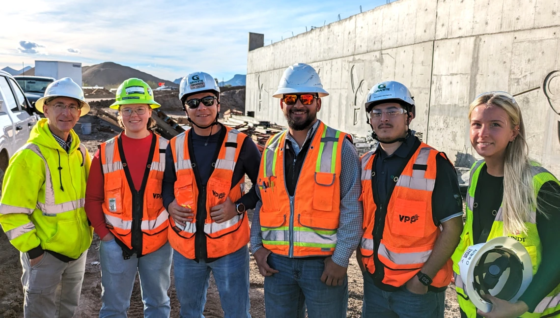 Six people pose outdoors at a construction site in hard hats