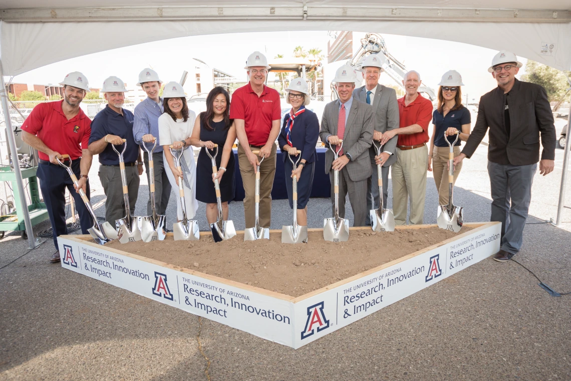 A group of a dozen people wearing white construction hats hold shovels that are placed into a square of dirt.