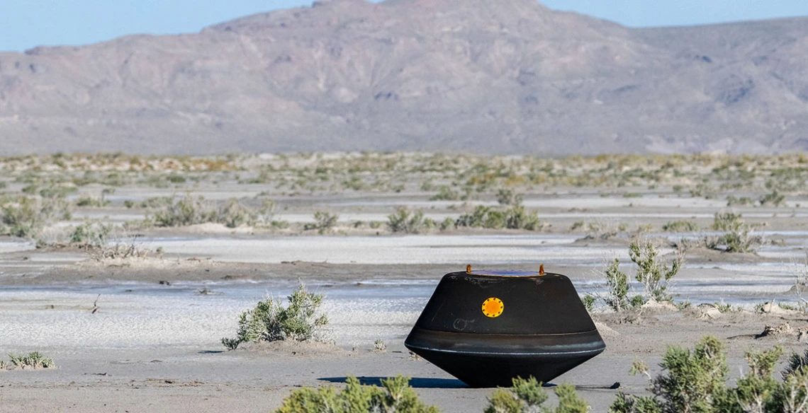 A black metal capsule the size of a doghouse rests on the desert ground