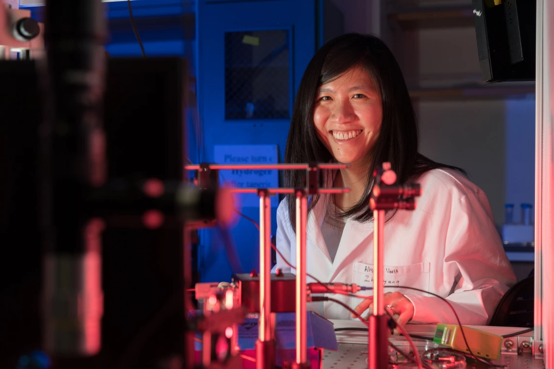 Judith Su wearing a white labcoat and smiling behind a complicated array of sensors.