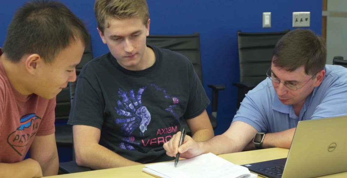 Three men lean over a sheet of paper and a laptop, the one on the far right pointing and explaining a concept.