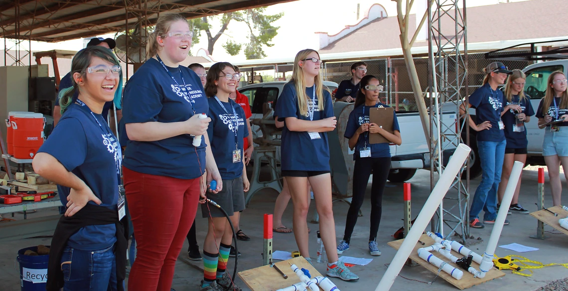 A group of high school girls wearing matching blue "Summer Engineering Academy" t-shirts grin off into the distance at a potato (off frame) they just launched out of a homemade potato launcher.