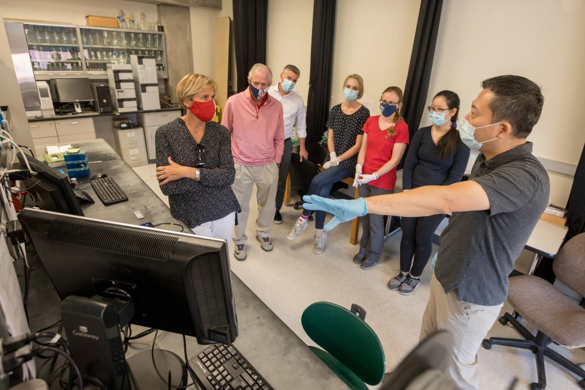 A man gestures to a computer on a laboratory counter, showing a small group of people something on the screen.