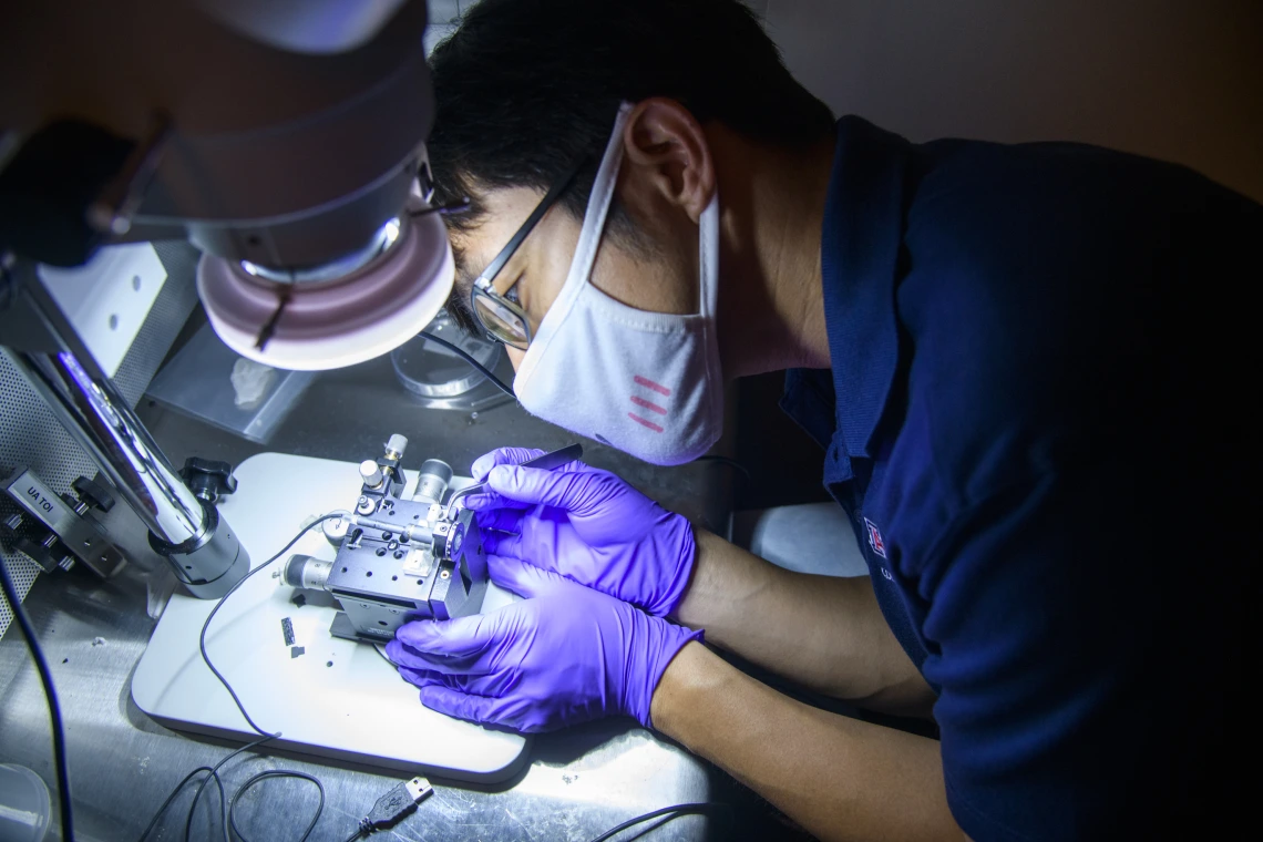 Dongkyun Kang, wearing rubber gloves and a mask, leans over to work on a small device under a bright light.