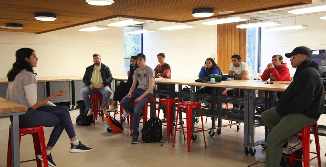 One student addresses a group of others in a room with tables and stools.