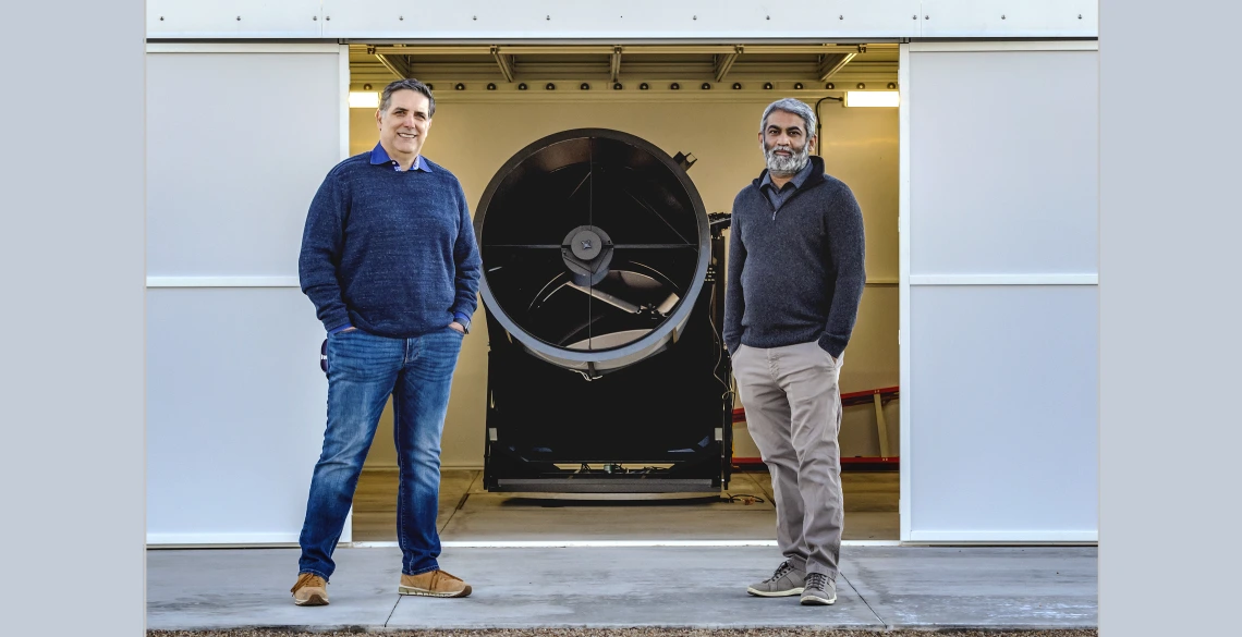 Roberto Furfaro and Vishnu Reddy stand in front of a piece of telescope equipment, a cylinder about 3-4 feet in diameter.