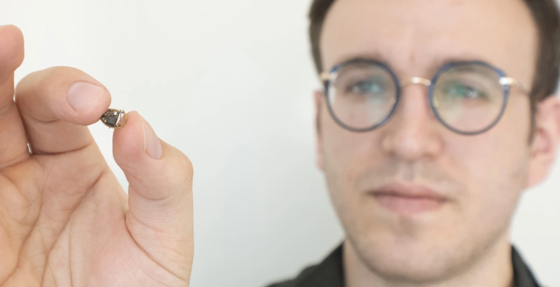 A man holds up a tiny electronic device, about the size of a chocolate chip.