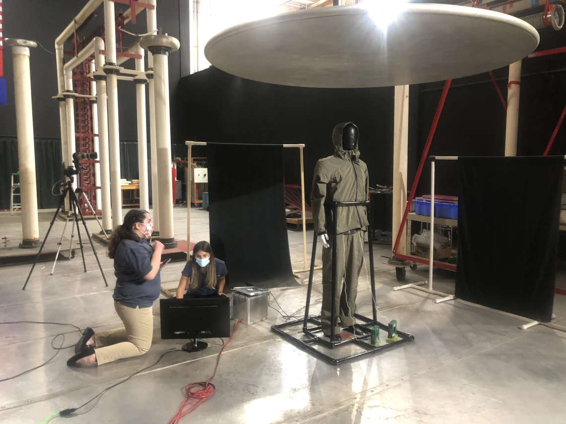 A mannequin covered in black tape is held up by a PVC pipe frame in a large room. Two students kneel next to electronic equipment near the mannequin.