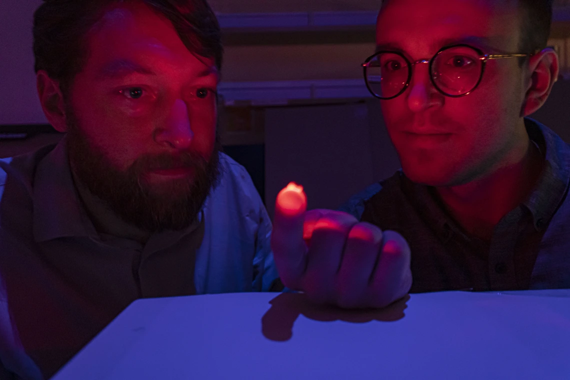 Two men, visible from the shoulders up, in a dimly lit room. The man on the left is holding a small, reddish light source on his hand, which is illuminating their faces. Both are staring at the light.