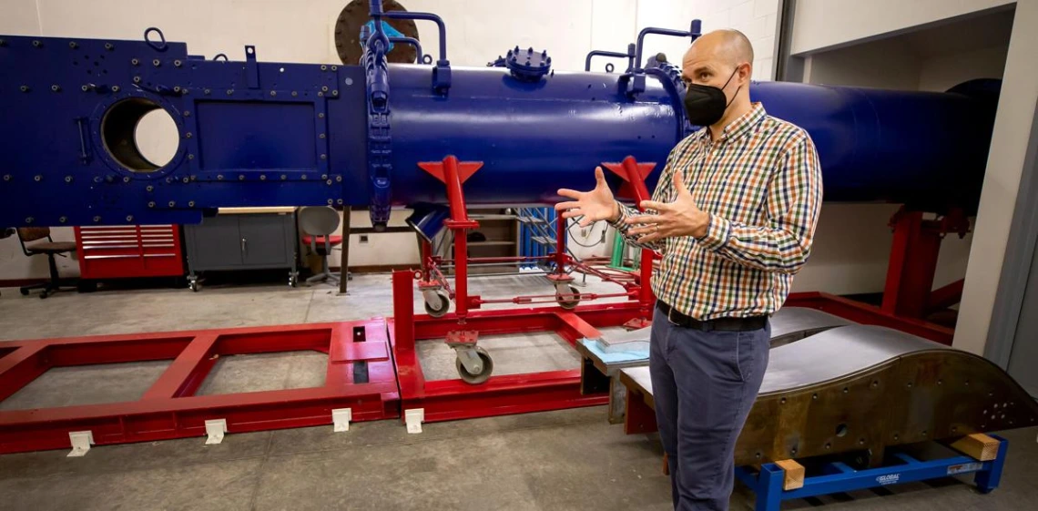 Jesse Little stands in front of a large blue wind tunnel -- it looks like a horizontal pipe -- on red scaffolding. He is wearing a black mask over his nose and mouth and gesturing with his hands to explain something.