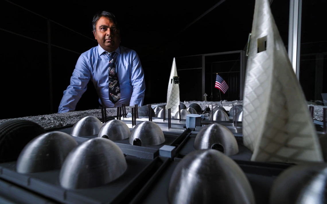 A man in a blue shirt stands in front of a small-scale table that mimics a lunar base.