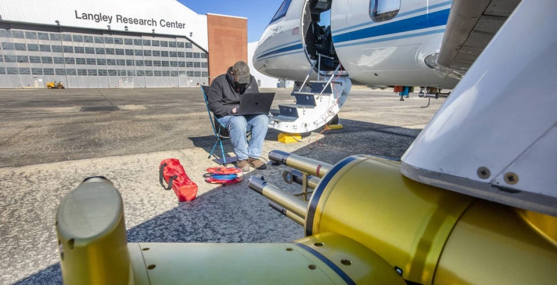 A man sits on a folding chair using a laptop, next to the steps of an airplane. The building behind him has a sign that says, "Langley Research Center."